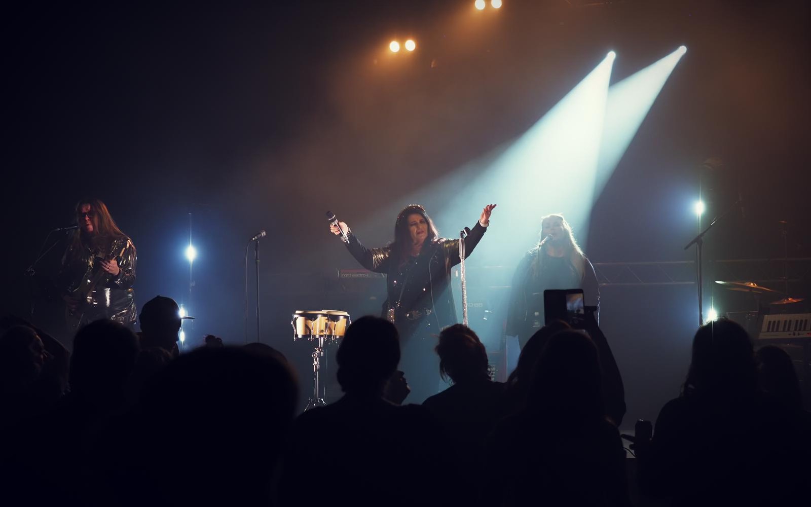 Meiju Suvas live on tour: the singer centre stage with hands raised, a guitarist to the left, cool white spotlight beams through haze behind and crowd silhouettes in the foreground