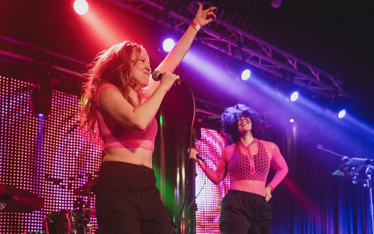 Two singers performing on stage: one sings into a handheld microphone with an arm raised, the other smiles with a mic in hand; a drum kit and colorful concert lighting behind them