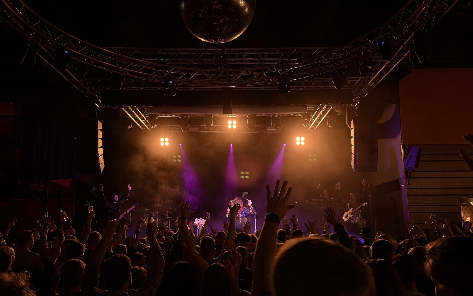 Meiju Suvas live on an indoor stage: the singer centre stage beneath a circular truss arch with a disco ball and warm and purple light beams, crowd hands raised in the foreground