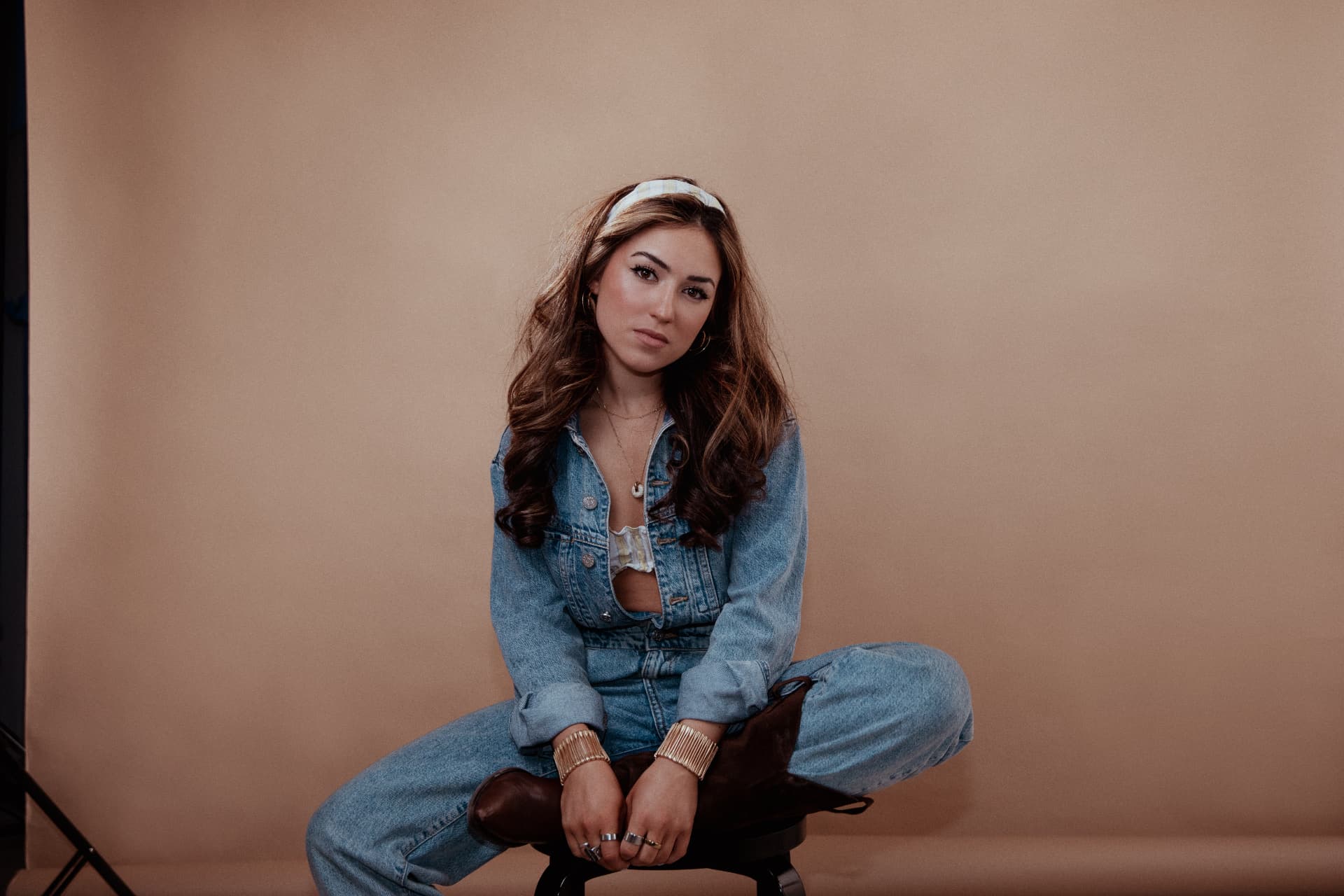 Ingrid in a studio portrait: the artist sits on a low stool in a light denim jacket and white headband against a warm beige backdrop