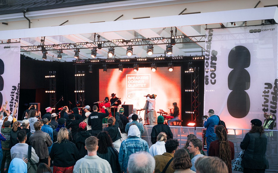 Oddfest outdoor stage: a band plays in front of a warm orange LED backdrop with the crowd watching from behind barriers